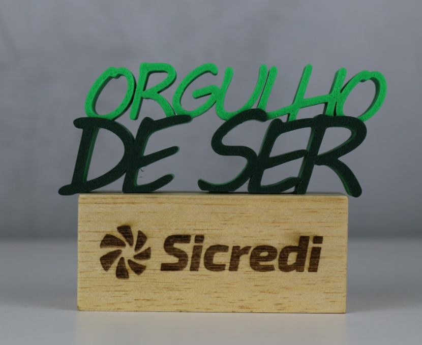 Troféu 'Orgulho de Ser' com letras 3D verdes sobre base de madeira com logo Sicredi